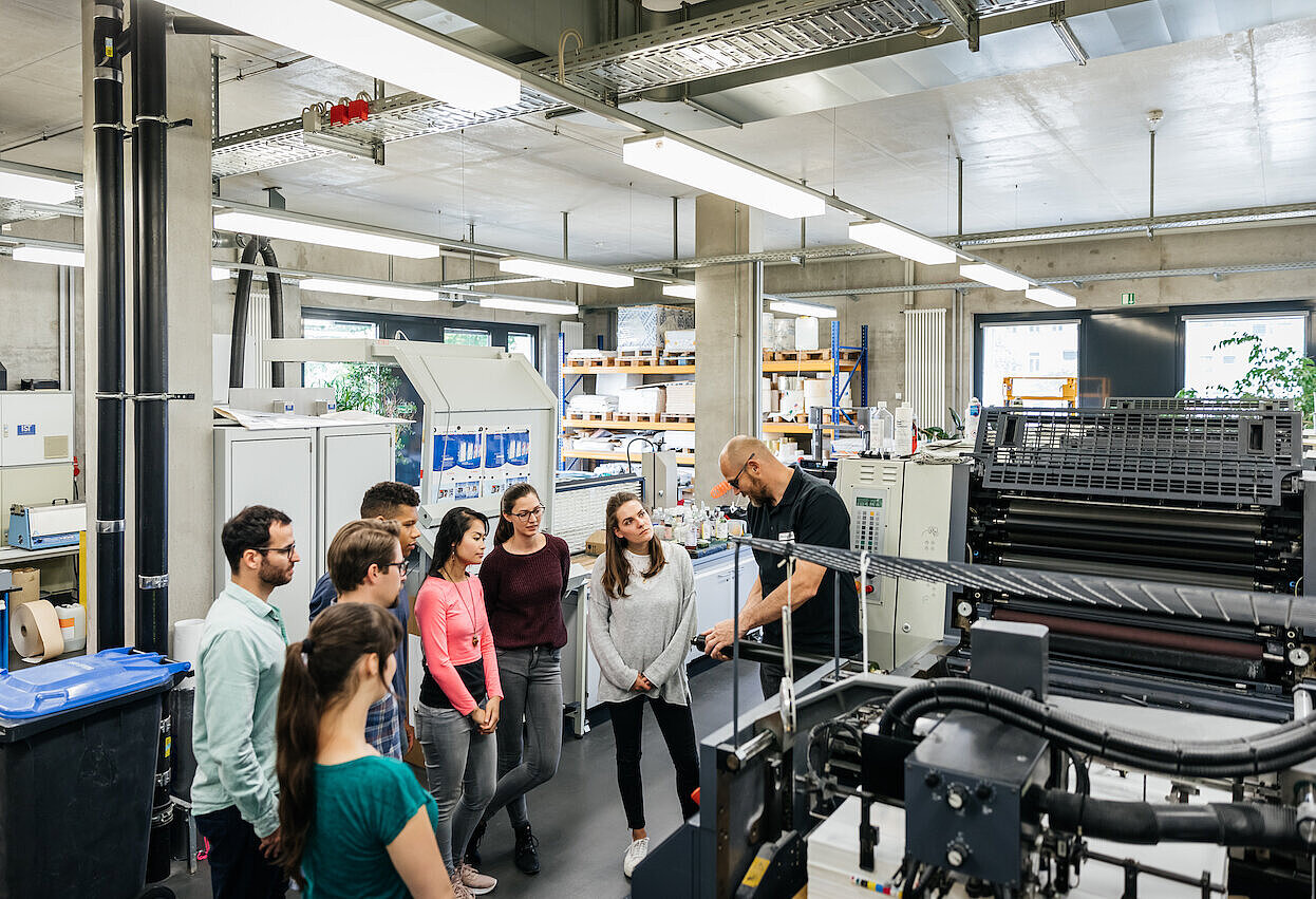 Gruppe Studierender bekommt eine Druckmaschine im Drucksaal erklärt.