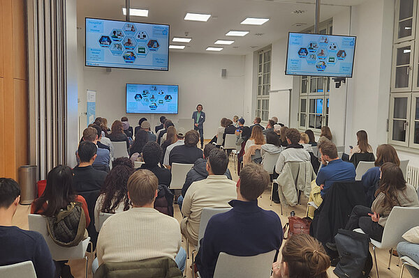 Ein länglicher großer Raum. Man sieht ca. 40 Studierende von hinten. Sie sitzen auf Stühlen und hören einem Unternehmensvertreter zu, der am Ende des Raums auf einem Podium steht und über sein Unternehmen spricht. Es sind auch drei Monitore zu sehen, auf denen denen seine Präsentation eingeblendet ist.